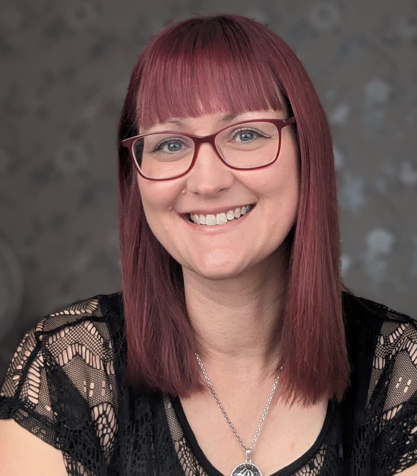 Photograph of Debbie Dawson. She is wearing a black top and Burgundy glasses and smiling at the camera.